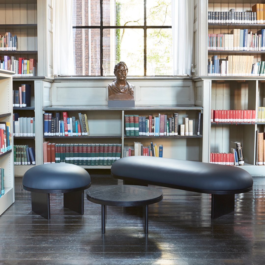 "Modern black bench and round table on dark wood floor, set against a backdrop of bookshelves."