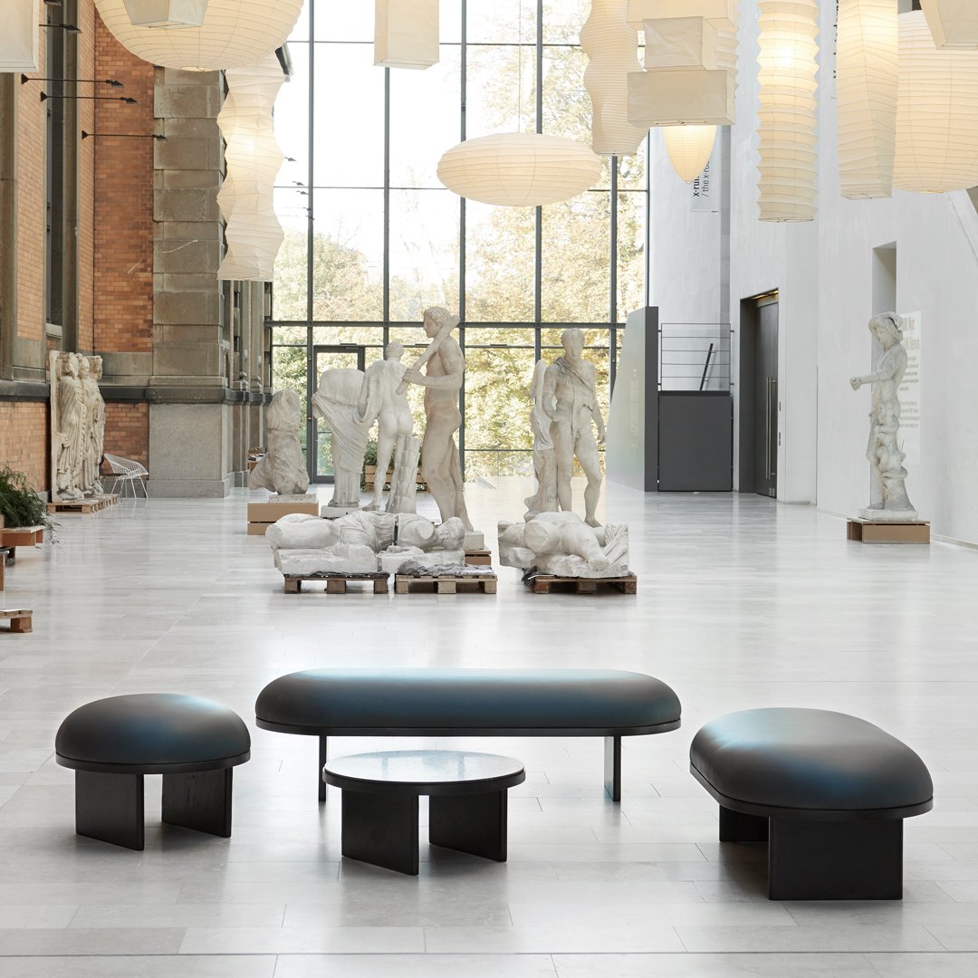 Modern black seating set with smooth curves and minimalistic design, enhanced by a neutral gallery backdrop and soft lighting.
