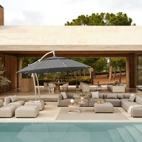 Gray umbrella over modern beige patio furniture by a pool, surrounded by green trees and natural wood accents.