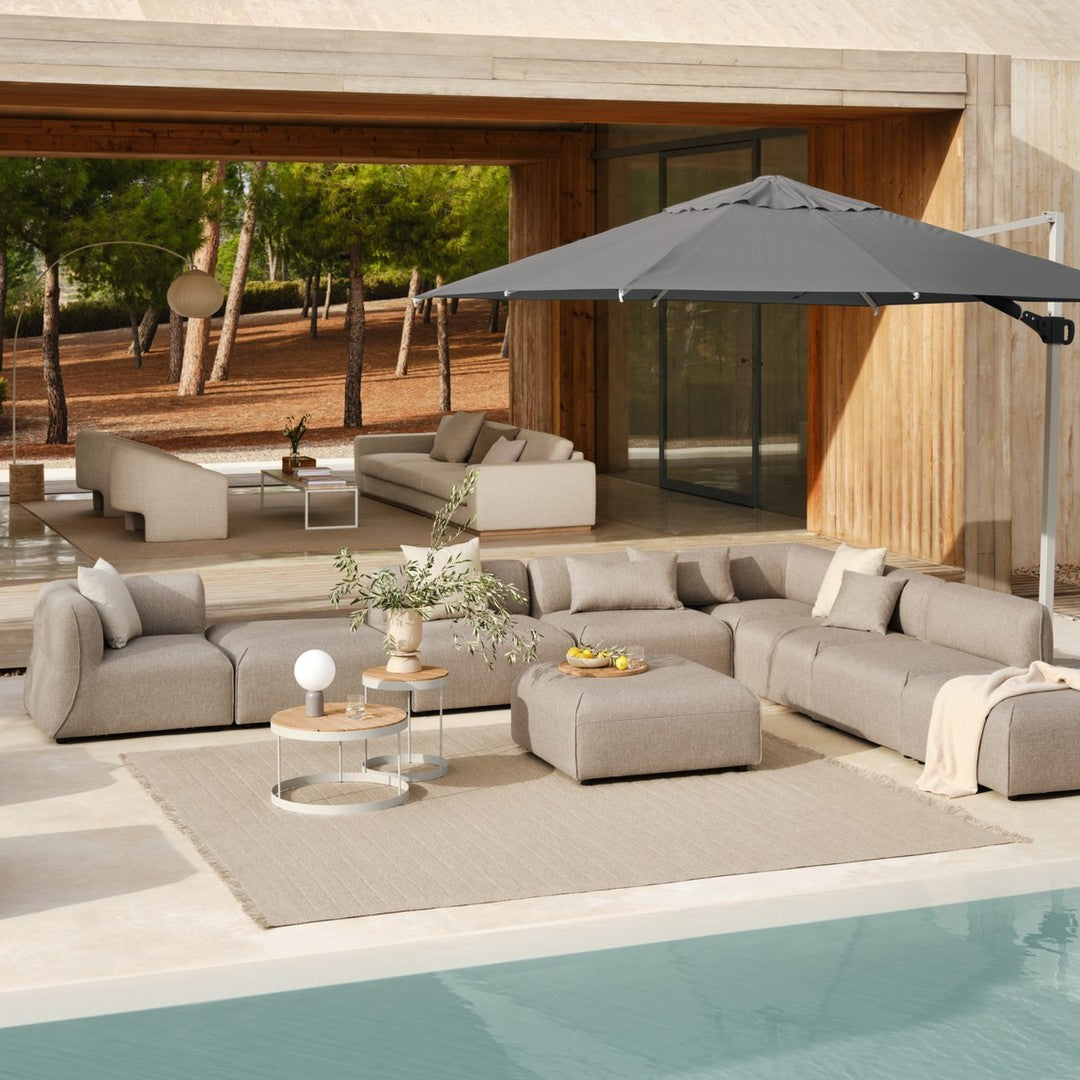 Cozy outdoor sectional sofa in light beige, with a dark gray umbrella and neutral accessories against a natural backdrop.