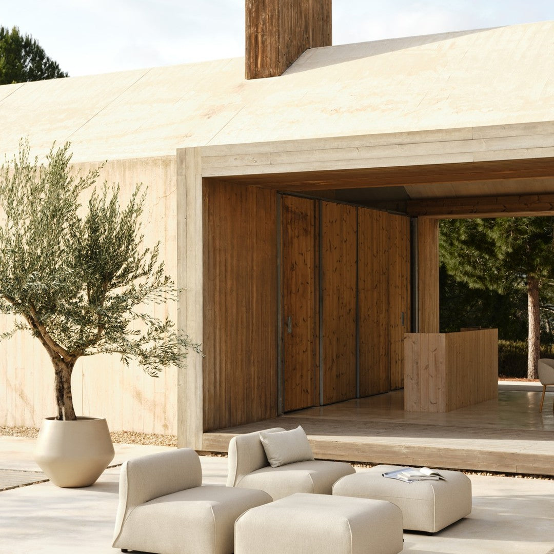 Modern neutral-colored furniture set against a natural wood and sandy background with olive greenery.
