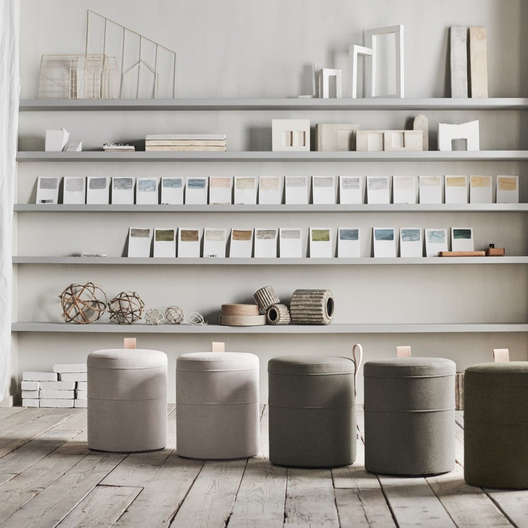 Neutral-colored round ottomans in light gray, beige, and olive green against a minimalist shelf backdrop.