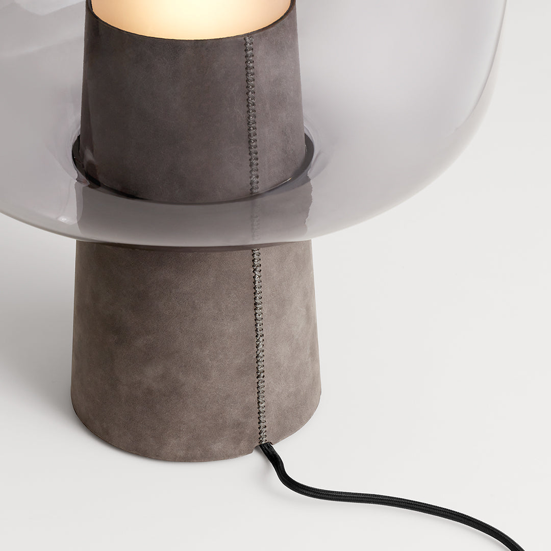 Gray suede lamp base with clear glass shade and black cord, featuring subtle stitching details for a modern look.