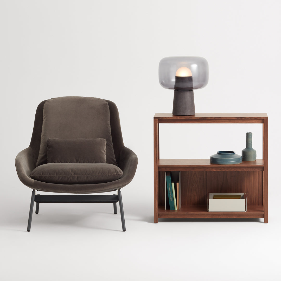 Brown upholstered chair and wooden shelf with teal and beige decor, illuminated lamp with a frosted glass shade.