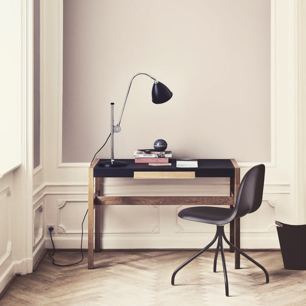 Modern wooden desk with a sleek black top and subtle beige wall backdrop, complemented by a dark chair and lamp.