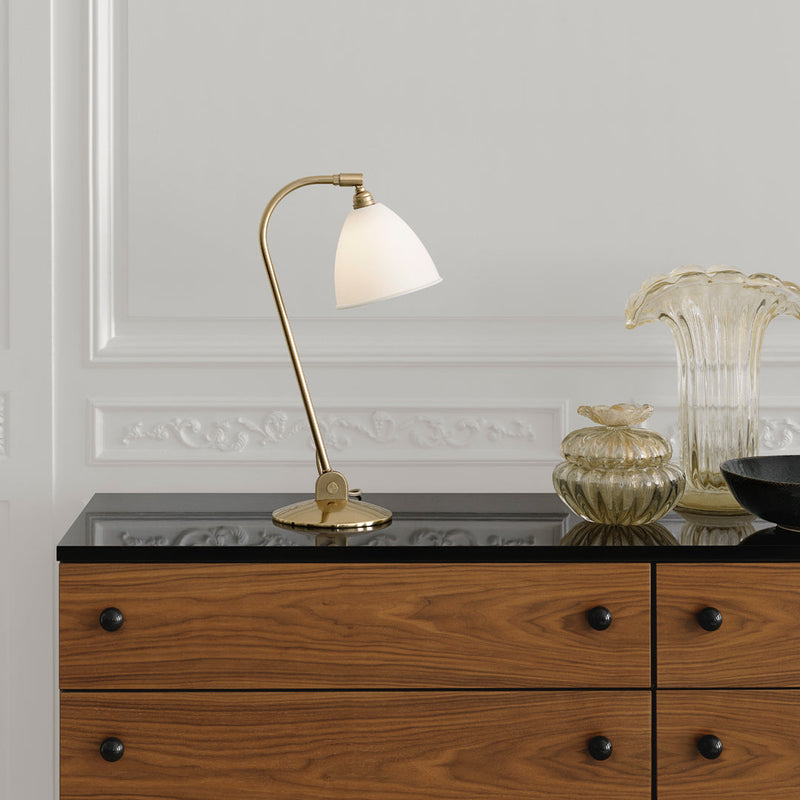 Elegant brass lamp with an opaque white shade, placed on a sleek black surface with a wooden chest of drawers.