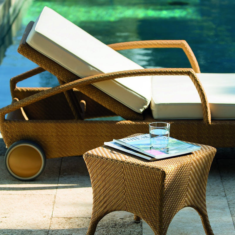 Woven rattan chaise lounge with cream cushions, paired with a textured brown side table, set by a turquoise pool.