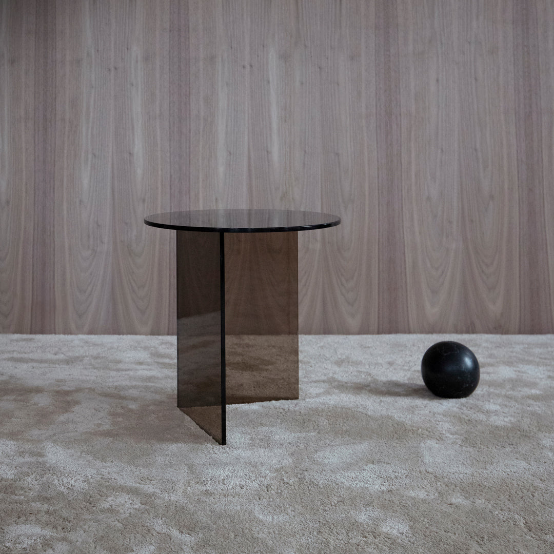Modern black glass side table with sleek geometric design, set against a textured beige carpet and wood panel backdrop.