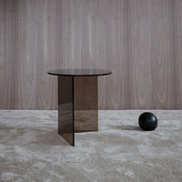 Modern black glass side table with sleek geometric design, set against a textured beige carpet and wood panel backdrop.