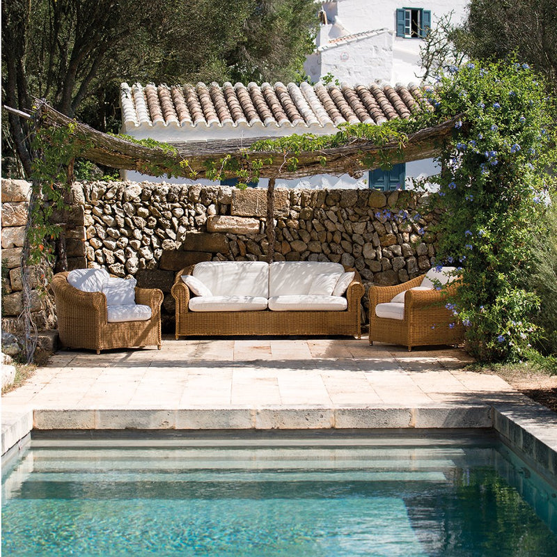 Warm brown wicker sofas with white cushions, surrounded by greenery and stone walls, next to a serene turquoise pool.