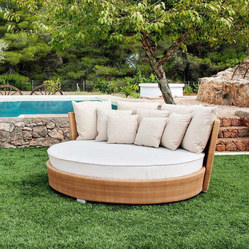 Round outdoor daybed with cream cushions and a natural wood finish, set on green grass near a pool.