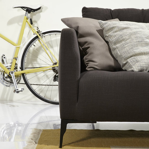Dark brown sofa with textured gray and beige pillows, complemented by a yellow bicycle in a bright, modern setting.