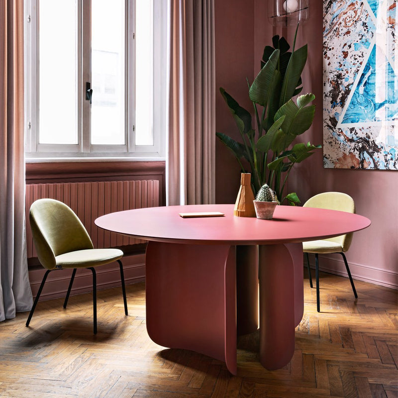 Round pink table with smooth surface, paired with green and beige chairs in a warm-toned, stylish interior setting.