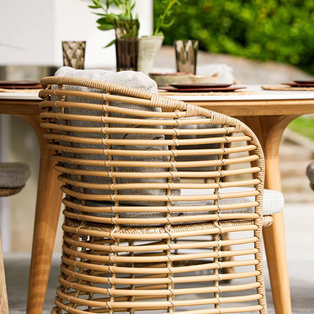 Natural rattan chair with a woven pattern, featuring a soft gray cushion against a bright outdoor setting.