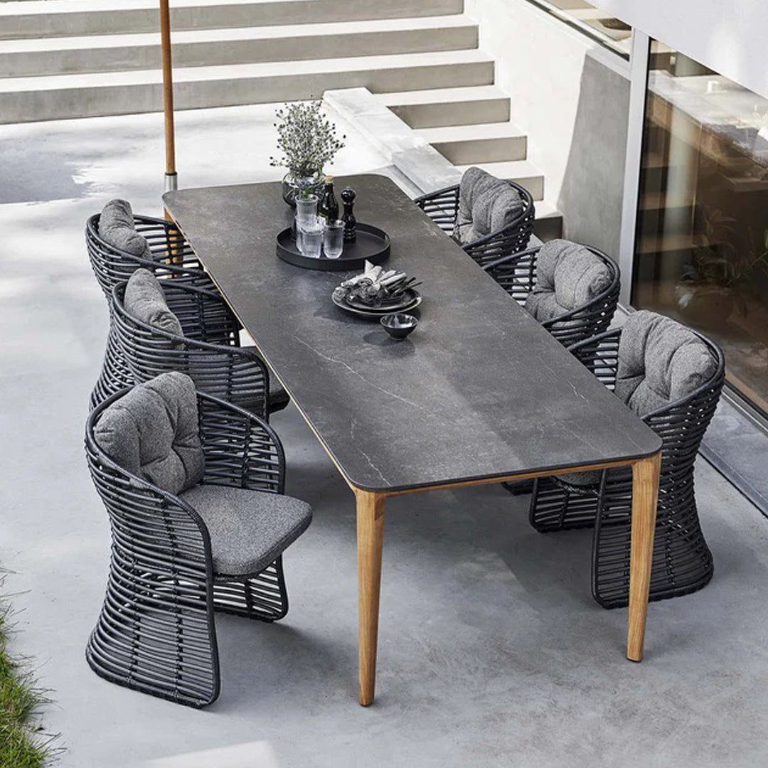 Long rectangular black stone table with light wood legs, surrounded by black woven chairs with gray cushions.