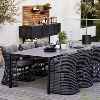 Black woven dining chairs surround a sleek black table, featuring a textured design and natural wood accents in the background.
