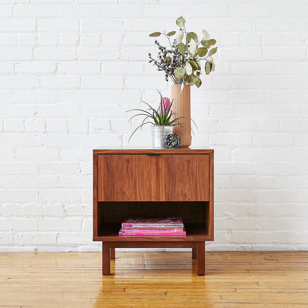 Brown wood nightstand with a minimalist design featuring smooth surfaces and a natural finish, complemented by green plants.