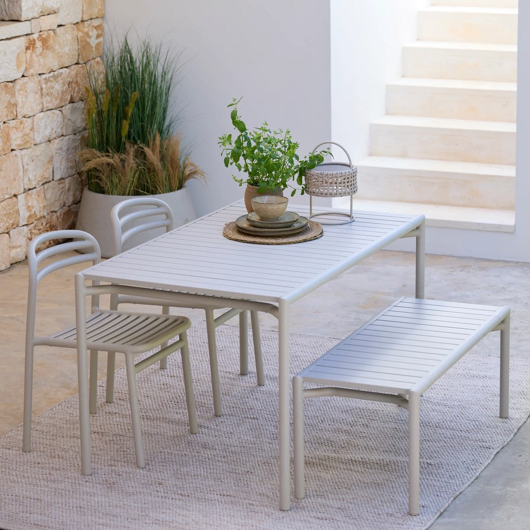 Light gray outdoor dining table set with slatted pattern and minimalistic design, surrounded by greenery.