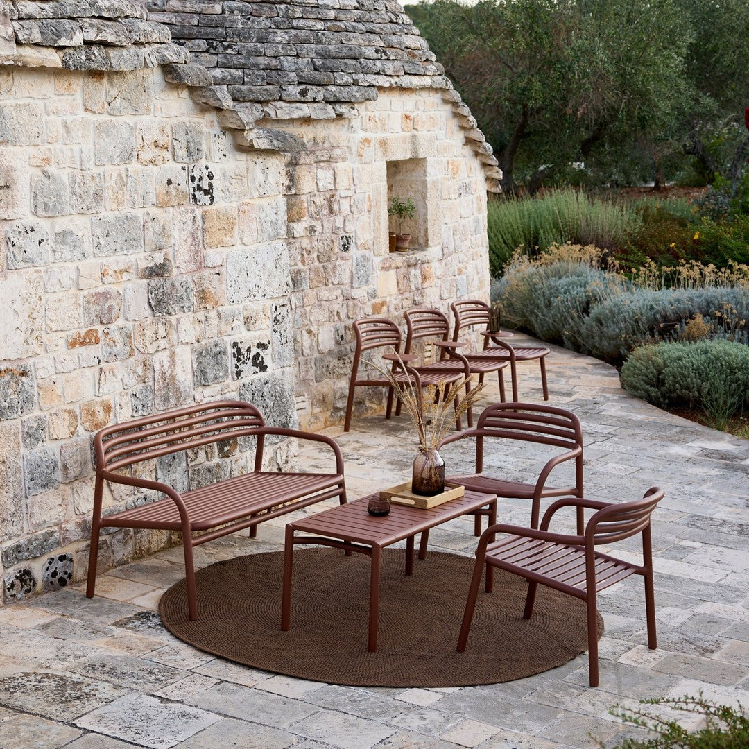 Brown metal outdoor furniture set with striped patterns on seats, paired with a round brown rug against a stone wall.