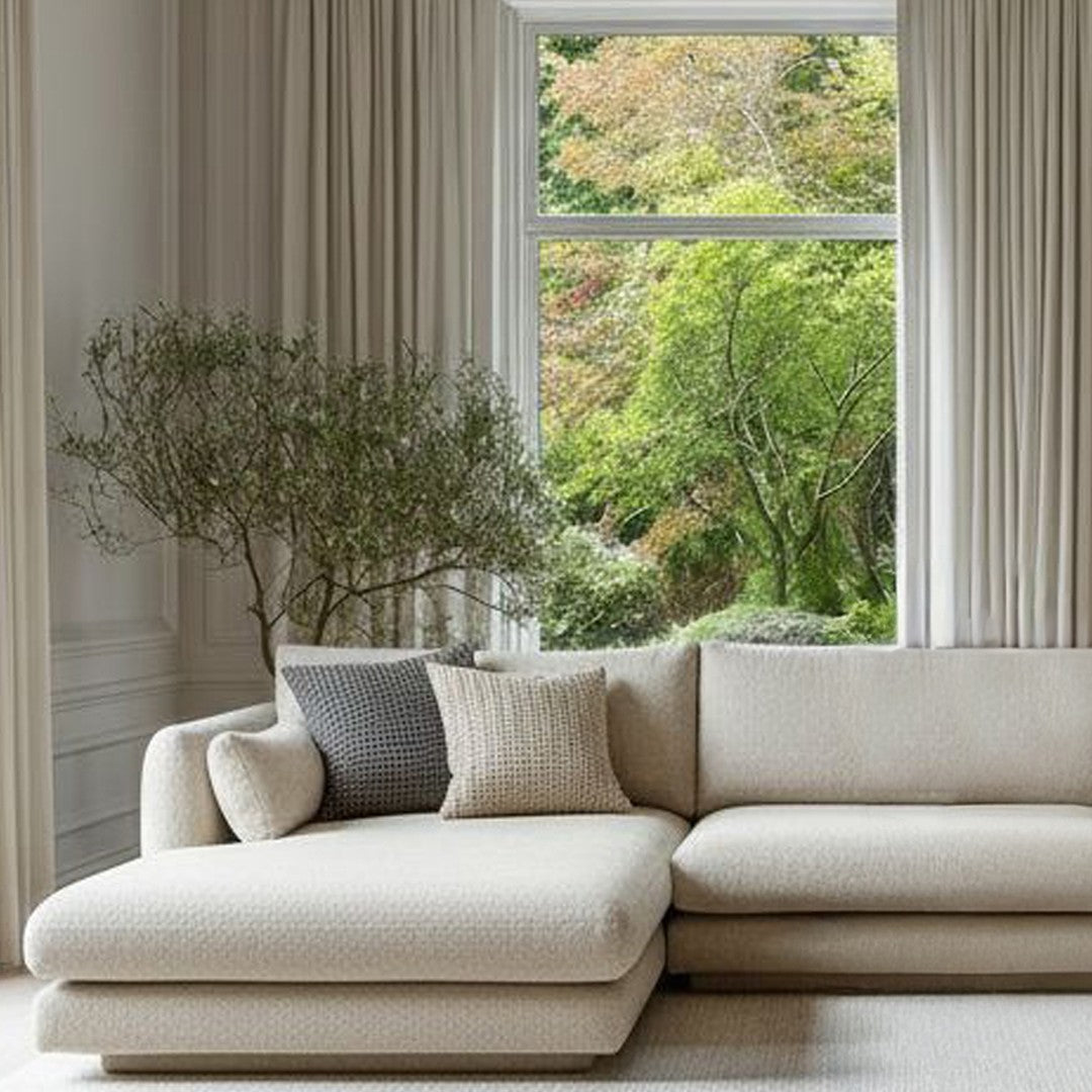 "Neutral beige sectional couch with textured throw pillows, complemented by soft light curtains and natural greenery outside."