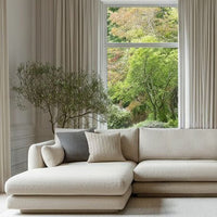 "Neutral beige sectional couch with textured throw pillows, complemented by soft light curtains and natural greenery outside."