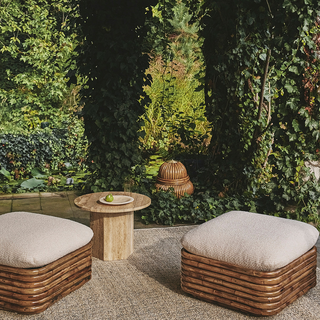"Natural woven rattan ottomans with light cream cushions, surrounded by lush greenery in a serene outdoor setting."