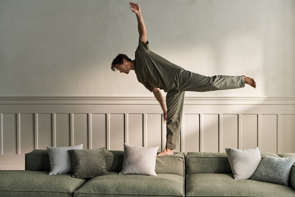 Person in green outfit performing a yoga pose on a green Cosima modular sofa by BOLIA with decorative pillows.