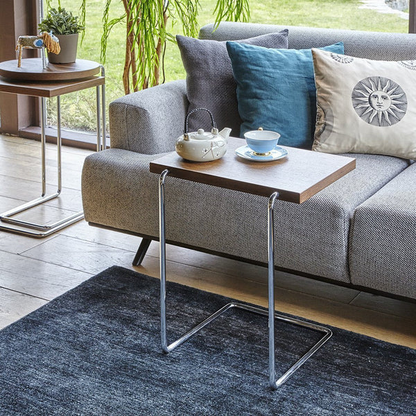 Modern side table with a walnut wood top and chrome legs, set against a gray sofa and patterned throw pillows.