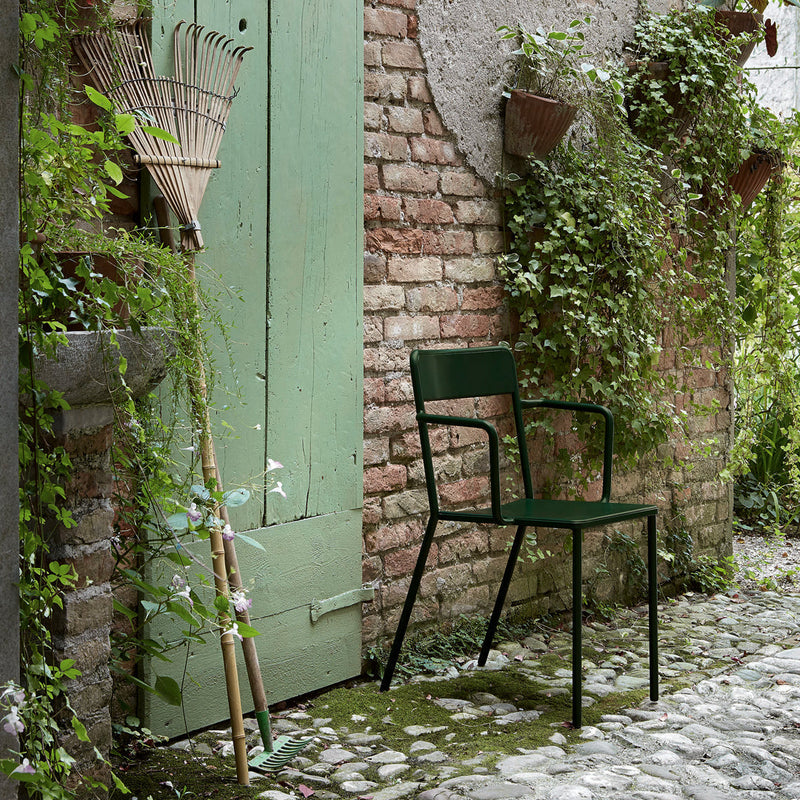 "Green metal chair against a brick wall, surrounded by lush greenery and a soft, natural color palette."