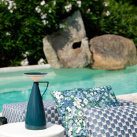 Teal vase, light pink rim, floral blue and green cushion, and checkered pattern in shades of blue and white by pool.