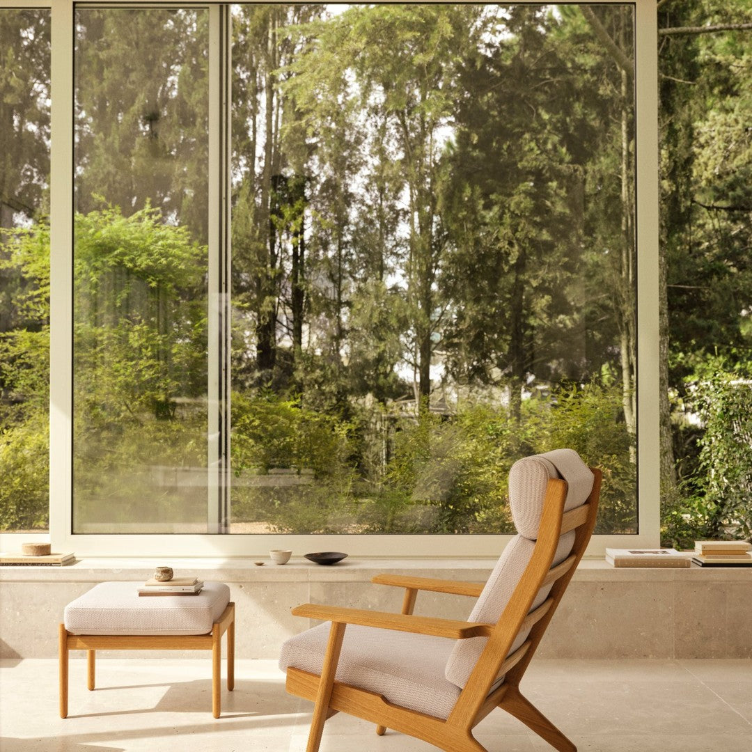 Modern wooden chair with beige cushions, framed by large windows showcasing lush green foliage outside.