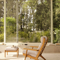 Modern wooden chair with beige cushions, framed by large windows showcasing lush green foliage outside.
