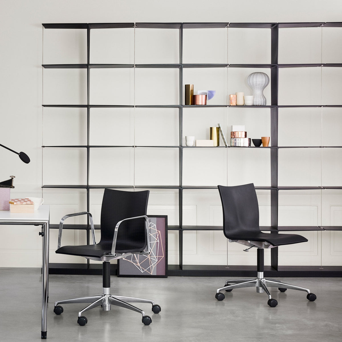Black ergonomic office chairs on a sleek gray floor, set against a minimalist white wall and geometric black shelving.