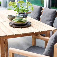 Wooden table with gray cushions, layered green and brown plates, and a black vase with green plants.