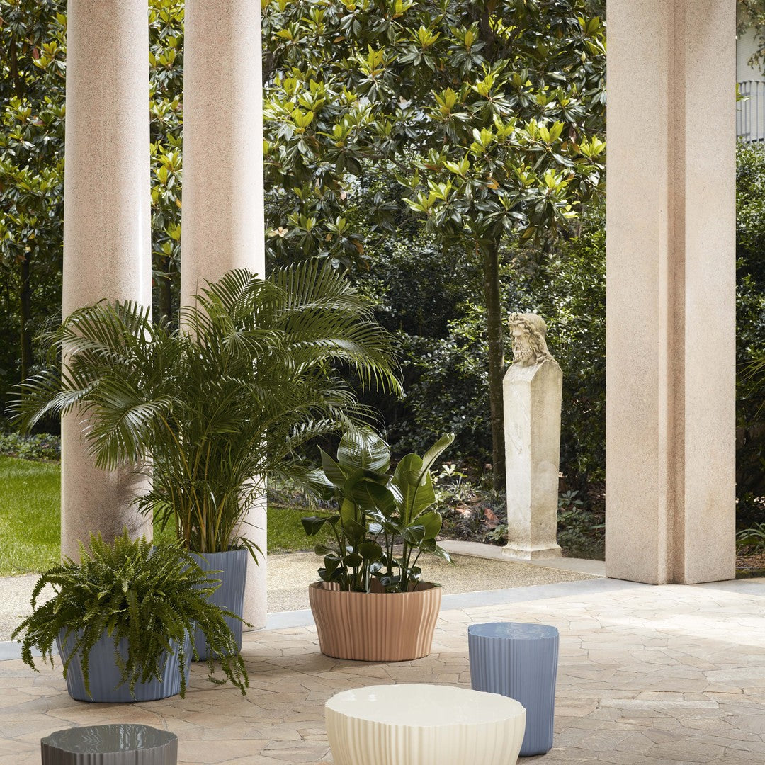 Stylish potted plants in muted blue, beige, and white tones, set against lush greenery and light stone pillars.