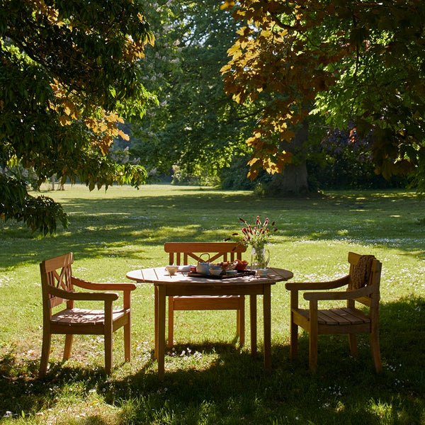 "Wooden dining set with natural finish, featuring a round table and two classic matching chairs surrounded by lush greenery."