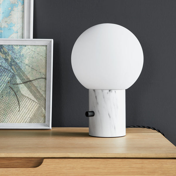 White globe lamp with a marble base, set against a dark wall and light wooden surface. Modern and sleek design.