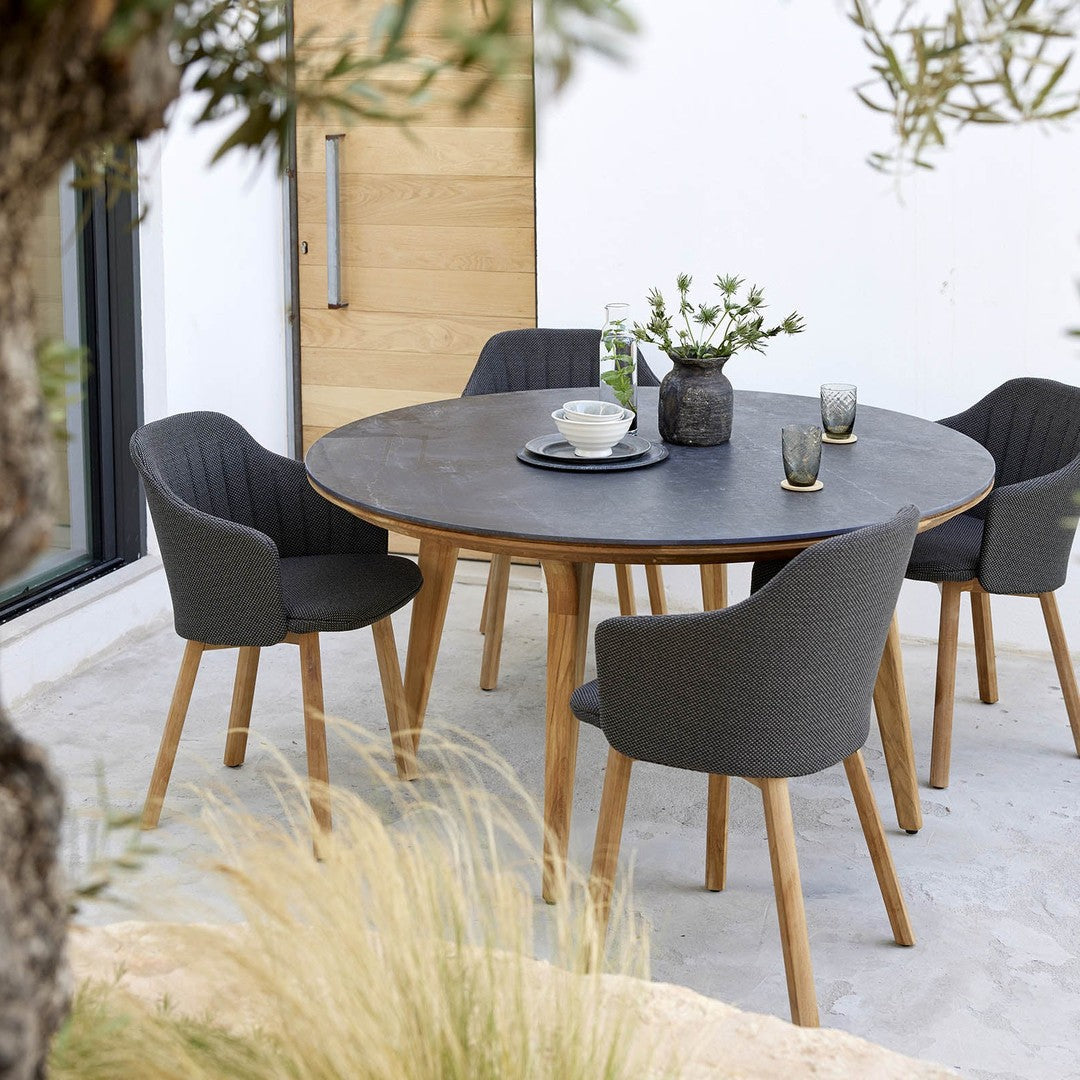 Round black stone dining table with gray upholstered chairs and wooden legs, set in a natural outdoor setting.
