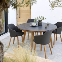 Round black stone dining table with gray upholstered chairs and wooden legs, set in a natural outdoor setting.