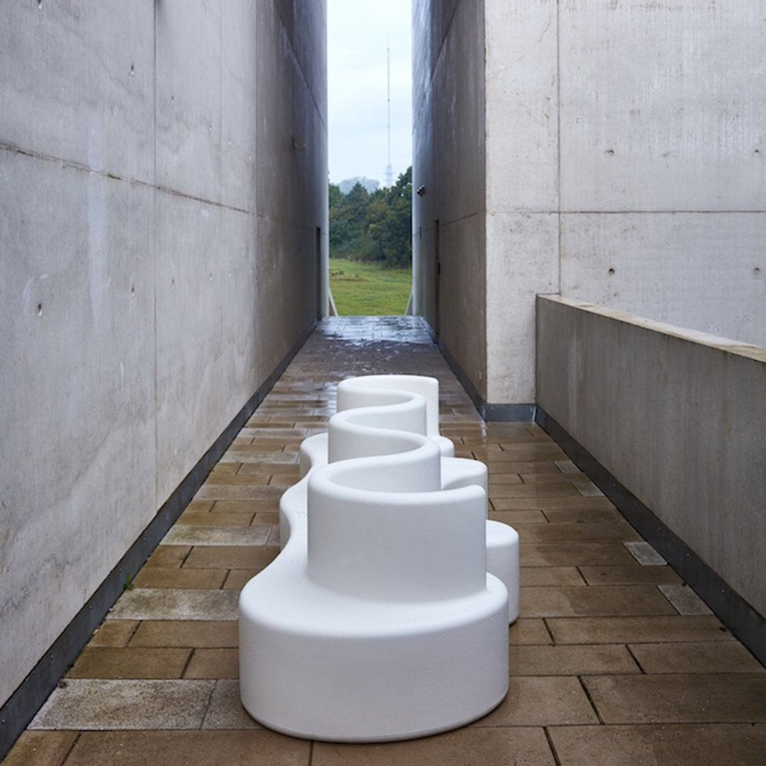 White abstract sculpture with a wavy design set against a gray concrete pathway and vibrant green landscape.