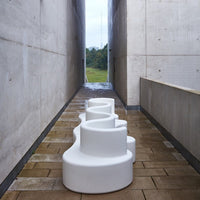 White abstract sculpture with a wavy design set against a gray concrete pathway and vibrant green landscape.