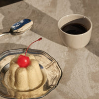 Yellow dessert with a red cherry on top, served on a silver plate, alongside a white cup of dark liquid.