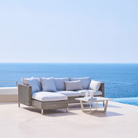 Gray woven outdoor sofa with light gray cushions, modern design, set against a clear blue sea and sky backdrop.