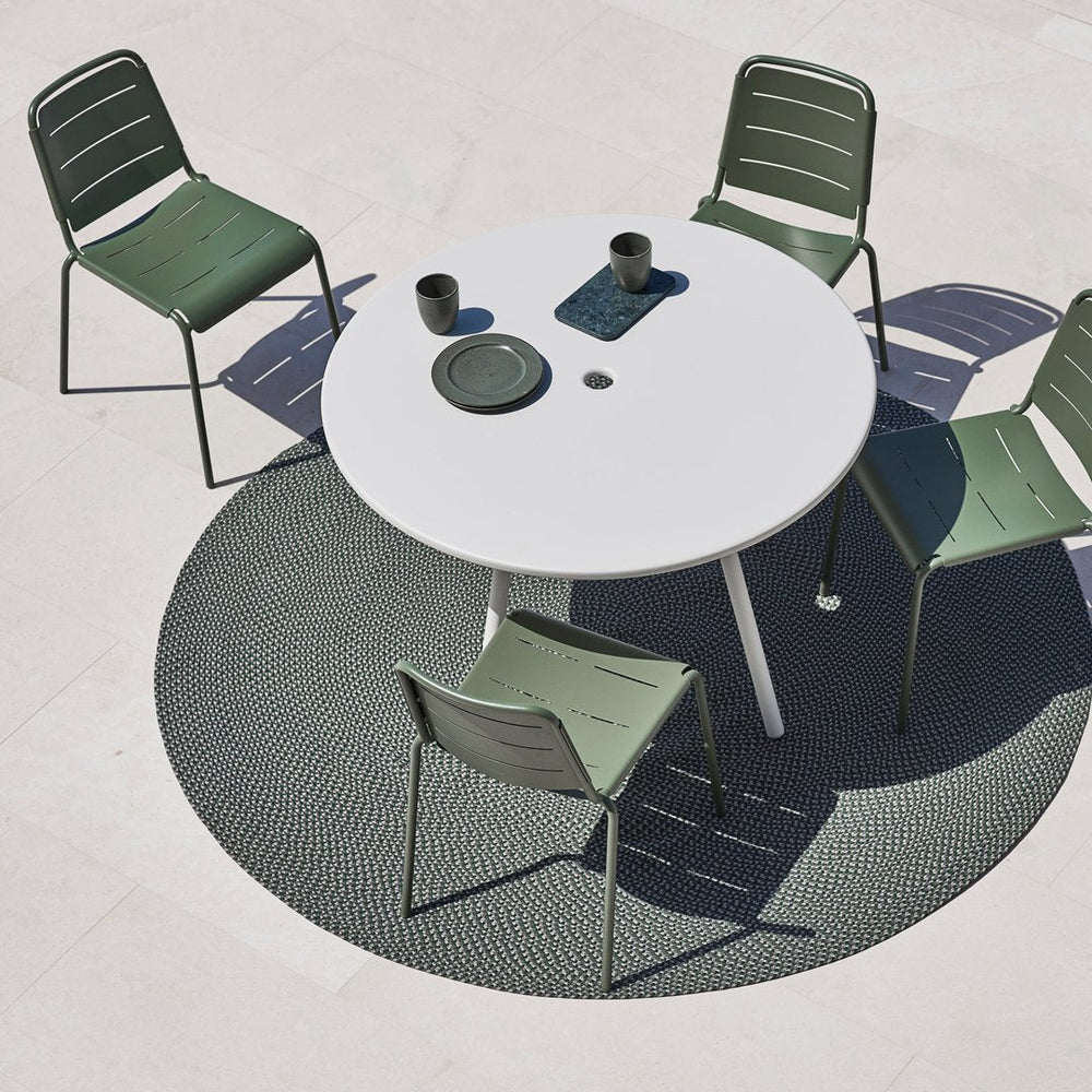 White round table with green metal chairs on a textured dark gray circular rug against a light marble floor.