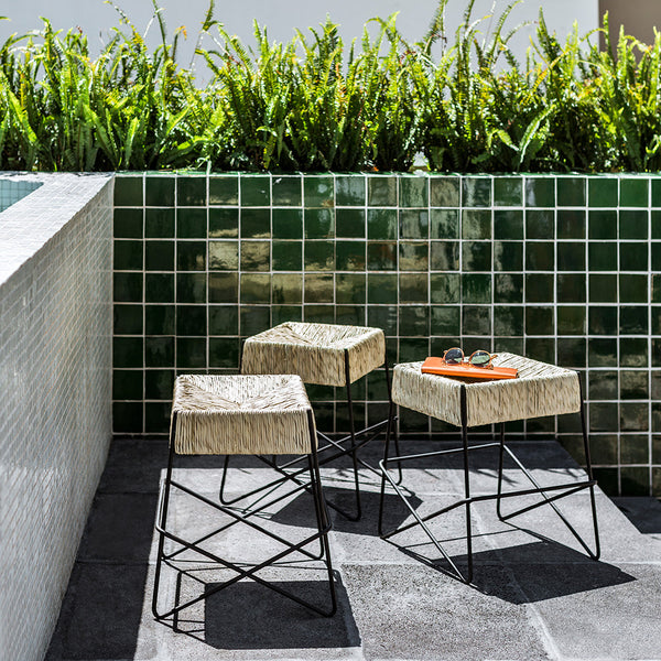 "Three modern stools with light beige tops and black metal frames, set against green tiled background and greenery."
