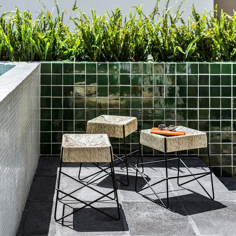 "Three modern stools with light beige tops and black metal frames, set against green tiled background and greenery."