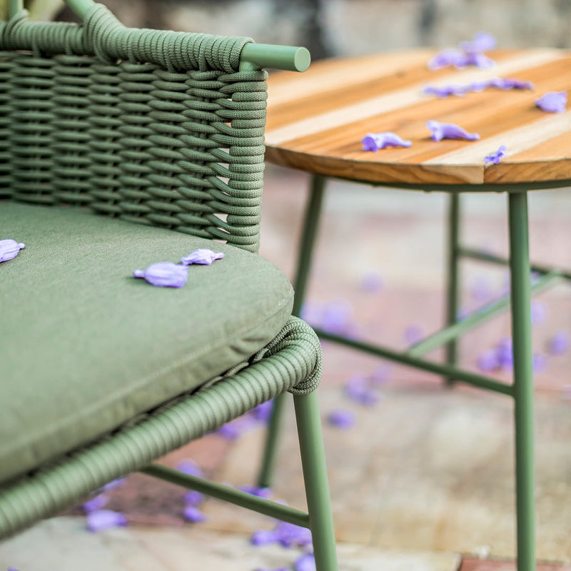 Green woven chair with olive cushions beside a natural wood table, accented with scattered purple petals.
