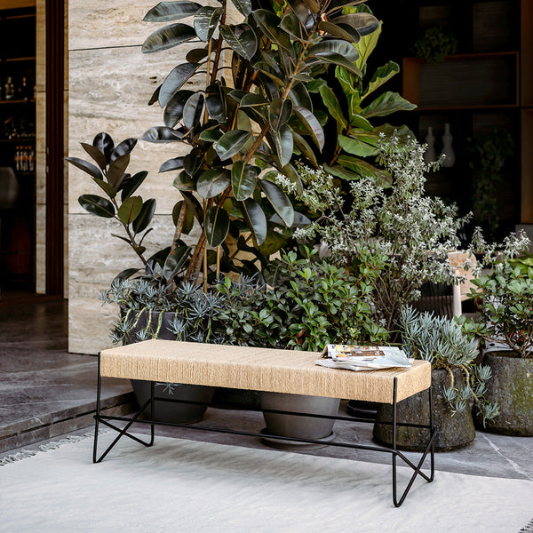 Natural wood bench with a textured beige surface and sleek black metal legs, surrounded by lush greenery.