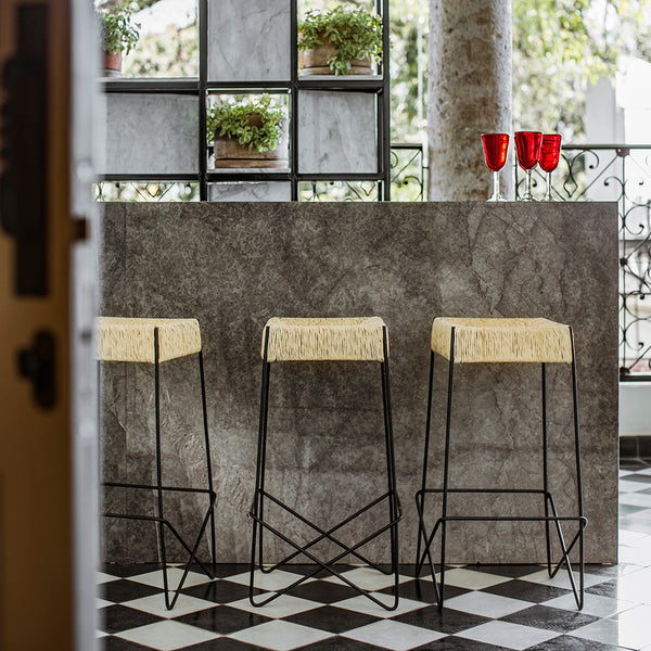 Black and white checkered floor with gray marble bar, featuring three straw-top stools and red glass decor.