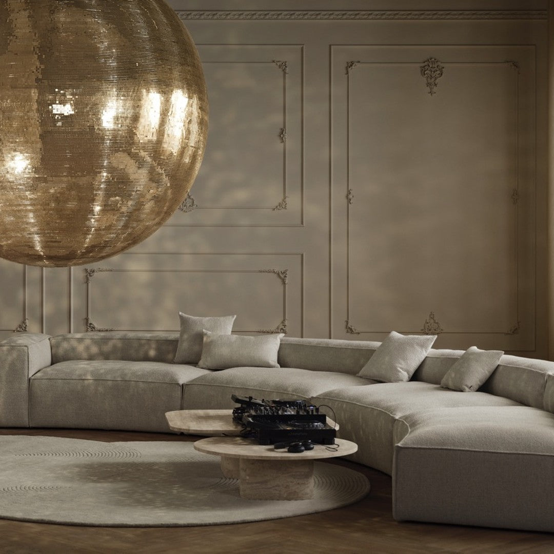 Curved beige sofa with textured pillows, round beige rug, and a shiny gold disco ball backdrop against soft beige walls.